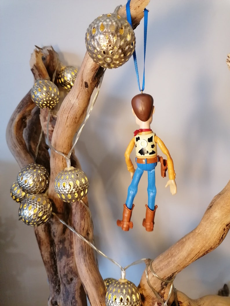 Puede incluir: Una rama de madera decorada con una cadena de luces y un adorno colgante de una figura de vaquero de juguete. Las luces son redondas y tienen un acabado met&aacute;lico con dise&ntilde;os recortados. La figura de vaquero lleva un chaleco marr&oacute;n, pantalones vaqueros azules y un sombrero marr&oacute;n.