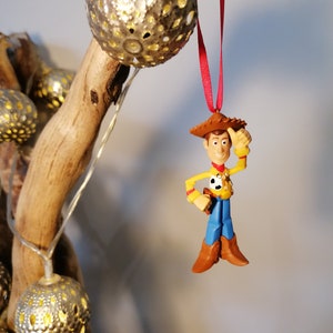 Pu&ograve; includere: Un filo rosso pende da un ramo con un ornamento dorato e una statuetta di plastica di Woody di Toy Story. Woody indossa il suo cappello da cowboy, il gilet e i jeans caratteristici.