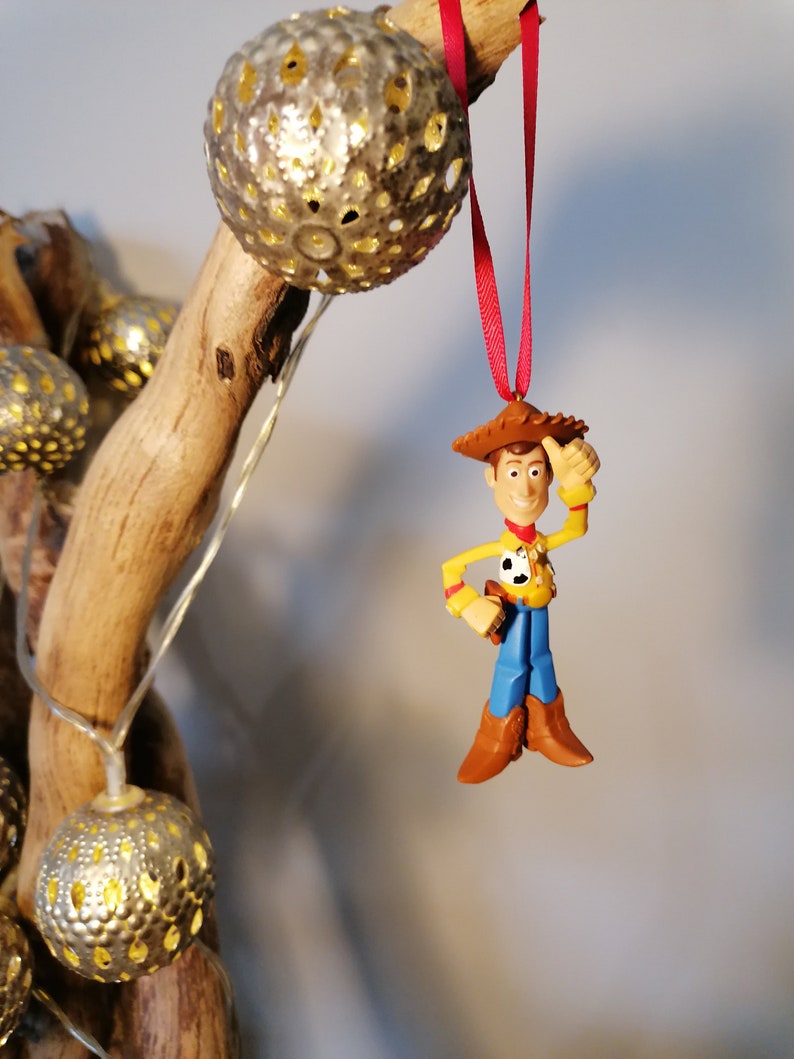 Pu&ograve; includere: Un filo rosso pende da un ramo con un ornamento dorato e una statuetta di plastica di Woody di Toy Story. Woody indossa il suo cappello da cowboy, il gilet e i jeans caratteristici.