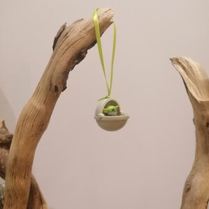 May include: A small, beige ornament featuring a green-skinned character in a floating pod, suspended by a light green ribbon. The ornament is hanging from a piece of driftwood, creating a whimsical display. The character has large eyes and a neutral expression.