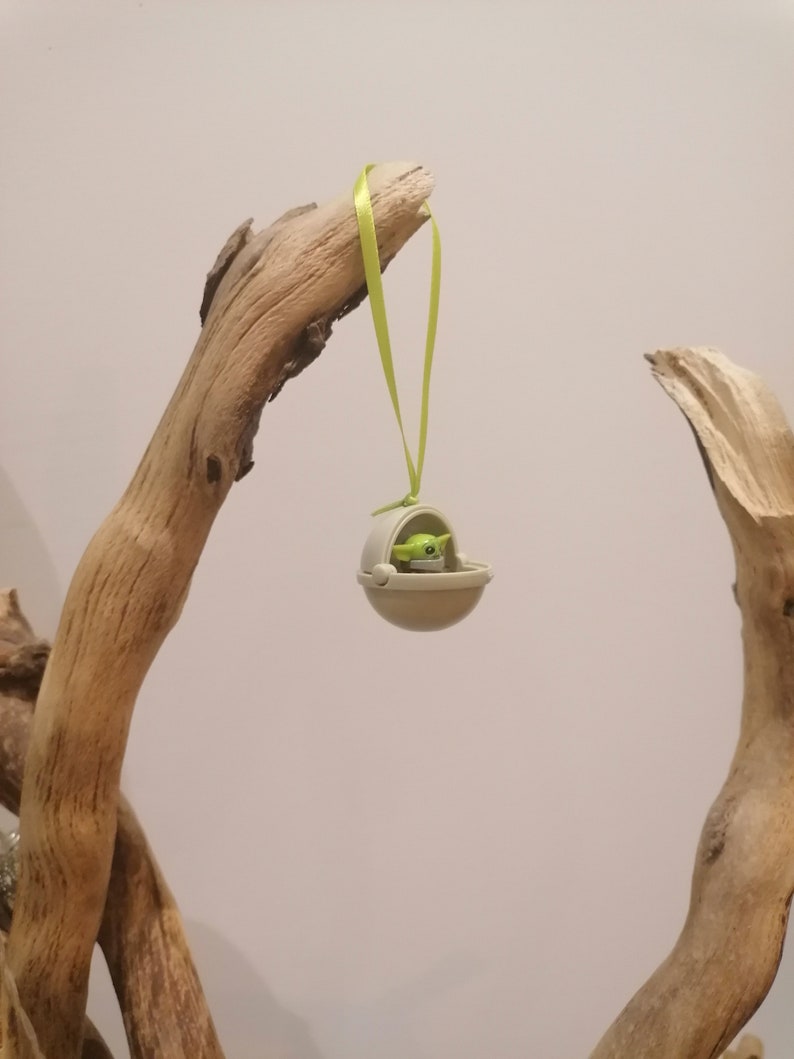 May include: A small, beige ornament featuring a green-skinned character in a floating pod, suspended by a light green ribbon. The ornament is hanging from a piece of driftwood, creating a whimsical display. The character has large eyes and a neutral expression.