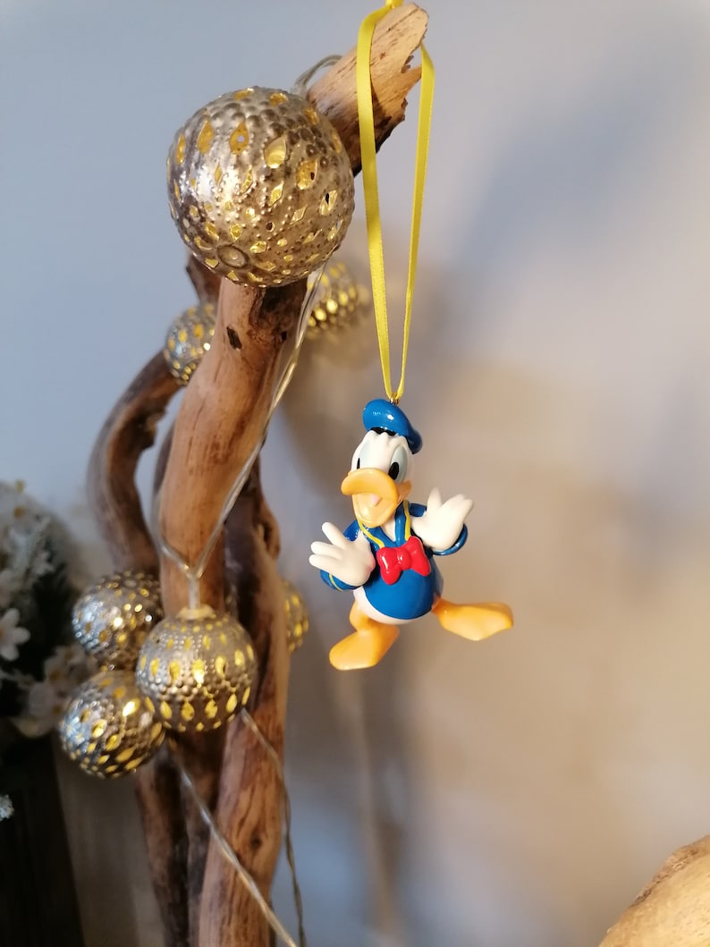 Op de afbeelding: Een gele draad hangt aan een stuk drijfhout met een blauw en geel Donald Duck ornament eraan. Het ornament is een kerstboomversiering.