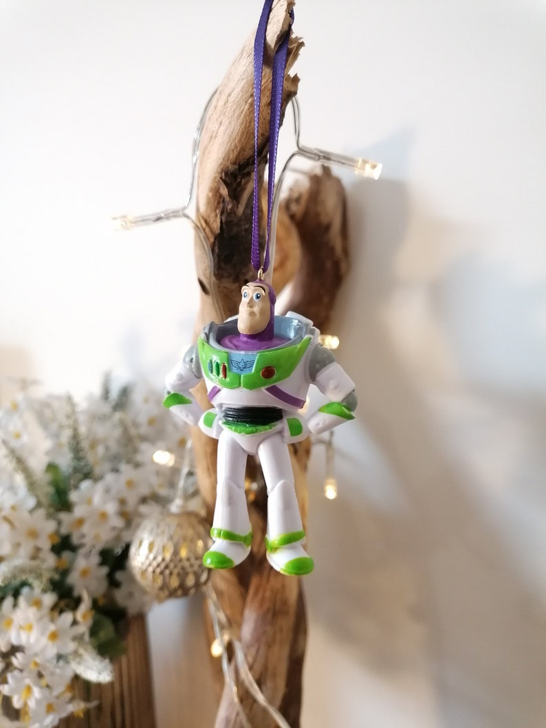 May include: A white and green plastic ornament of Buzz Lightyear from the Toy Story franchise. The ornament is hanging from a purple ribbon.