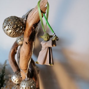 Puede incluir: Adorno navide&ntilde;o de Yoda con un sable de luz verde, colgado de una cinta verde. La figura de Yoda lleva una t&uacute;nica beige. El adorno se exhibe en un trozo de madera flotante, con bolas plateadas y luces de hadas.