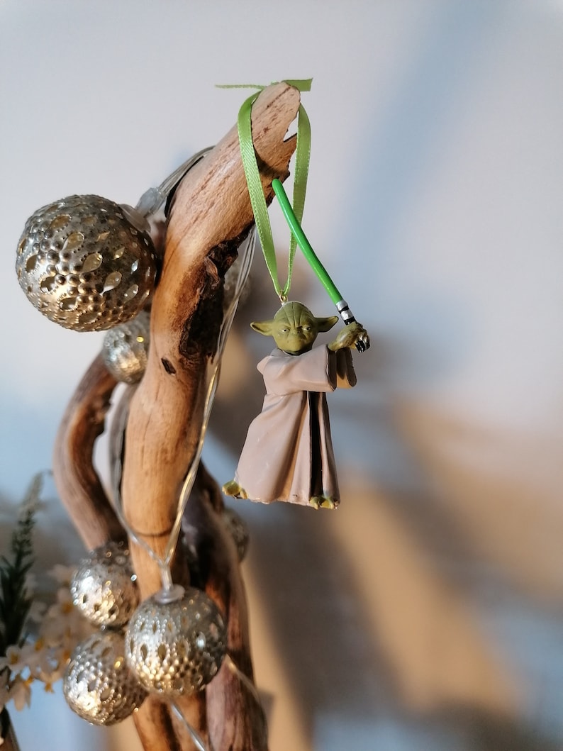 Puede incluir: Adorno navide&ntilde;o de Yoda con un sable de luz verde, colgado de una cinta verde. La figura de Yoda lleva una t&uacute;nica beige. El adorno se exhibe en un trozo de madera flotante, con bolas plateadas y luces de hadas.