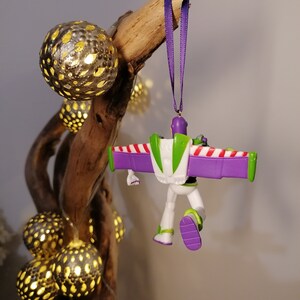 May include: Purple and green Buzz Lightyear Christmas ornament with candy cane wings hanging from a purple ribbon on a branch with gold light bulbs.
