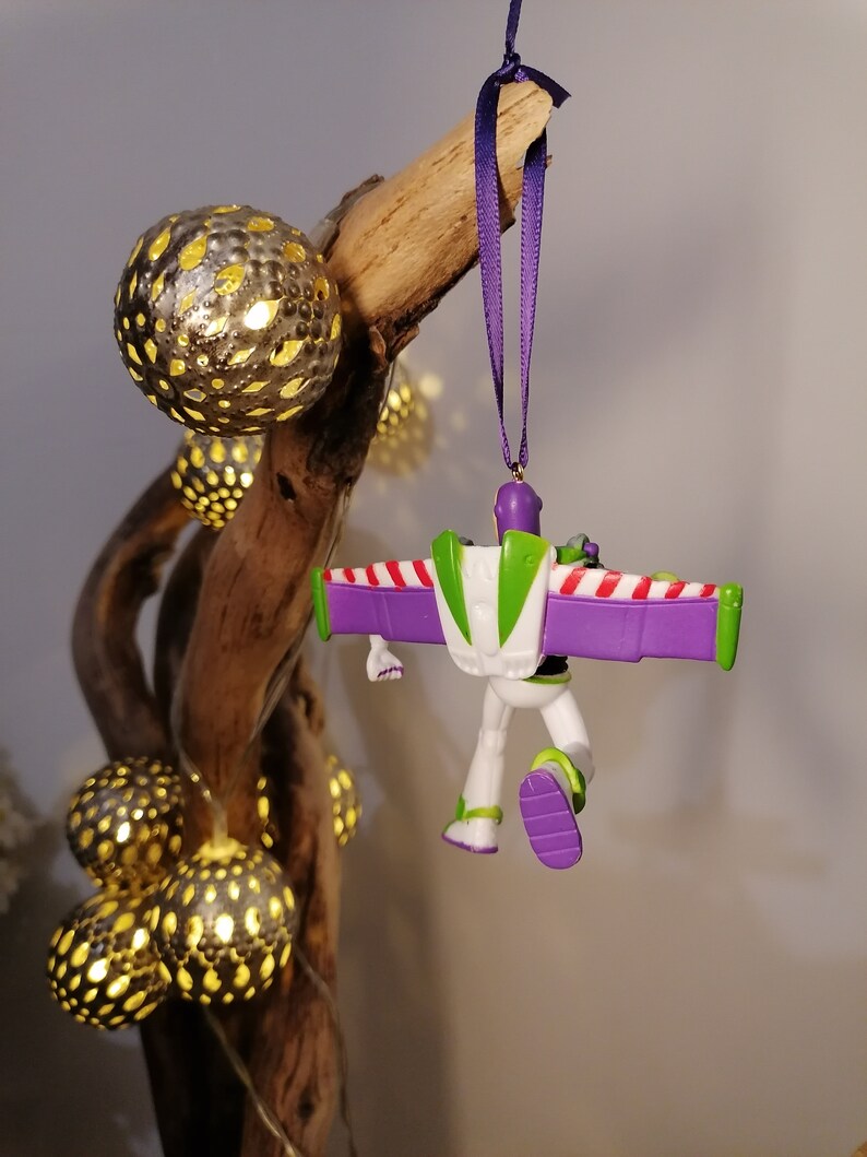 May include: Purple and green Buzz Lightyear Christmas ornament with candy cane wings hanging from a purple ribbon on a branch with gold light bulbs.
