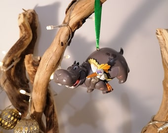 May include: A gray hippopotamus ornament with a yellow and orange bird perched on its back. The ornament is hanging from a green ribbon on a branch with white lights.