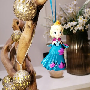 K&ouml;nnte beinhalten: Ein blauer und lila Prinzessinnen-Anh&auml;nger, der an einem Ast mit goldenen Lichterketten h&auml;ngt. Die Puppe tr&auml;gt ein blaues Kleid mit lila Akzenten und eine Krone.