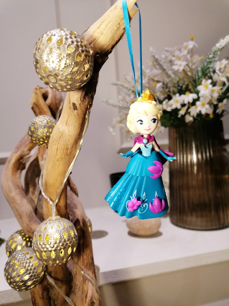 K&ouml;nnte beinhalten: Ein blauer und lila Prinzessinnen-Anh&auml;nger, der an einem Ast mit goldenen Lichterketten h&auml;ngt. Die Puppe tr&auml;gt ein blaues Kleid mit lila Akzenten und eine Krone.