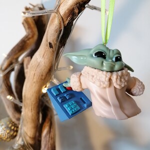 May include: A green and brown Yoda-like character ornament with a pink coat and a blue control panel in its hand. The ornament is hanging from a green ribbon on a wooden branch.