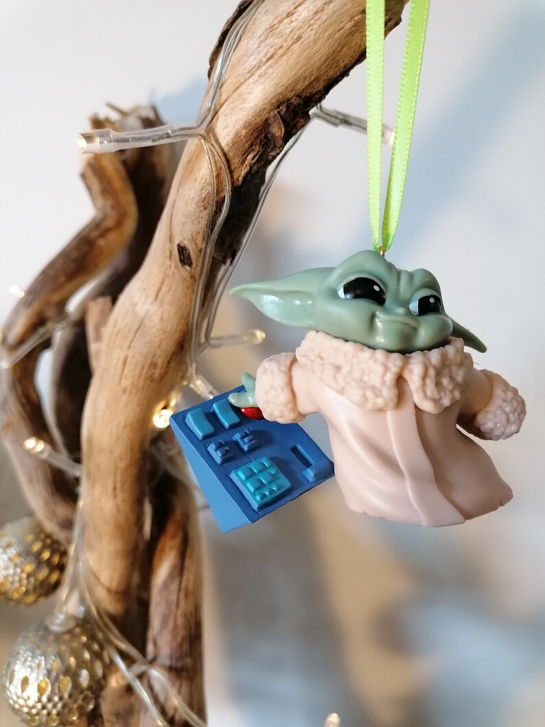 May include: A green and brown Yoda-like character ornament with a pink coat and a blue control panel in its hand. The ornament is hanging from a green ribbon on a wooden branch.
