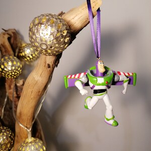 May include: A purple and green plastic toy ornament of Buzz Lightyear from the Toy Story franchise. The ornament is hanging from a purple ribbon on a branch with gold decorative balls.