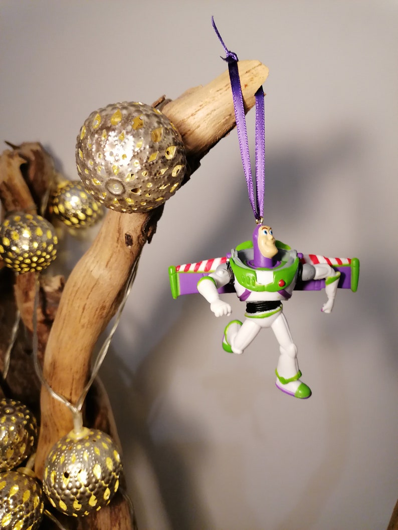 May include: A purple and green plastic toy ornament of Buzz Lightyear from the Toy Story franchise. The ornament is hanging from a purple ribbon on a branch with gold decorative balls.