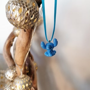 May include: A blue Stitch character charm hangs from a turquoise ribbon necklace. The charm is a 3D representation of the popular Disney character.