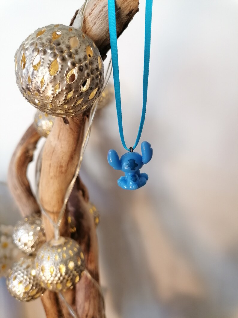 May include: A blue Stitch character charm hangs from a turquoise ribbon necklace. The charm is a 3D representation of the popular Disney character.