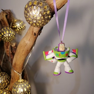 May include: A purple and white Buzz Lightyear Christmas ornament hanging from a branch with a purple ribbon. The ornament is a plastic figurine of Buzz Lightyear from the Toy Story movie franchise.