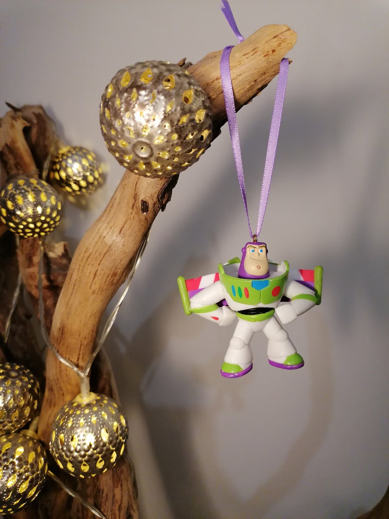May include: A purple and white Buzz Lightyear Christmas ornament hanging from a branch with a purple ribbon. The ornament is a plastic figurine of Buzz Lightyear from the Toy Story movie franchise.