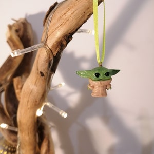 May include: A green and tan Baby Yoda ornament hanging from a light green ribbon. The ornament is suspended from a piece of driftwood, with a string of small white lights wrapped around it. The background is a plain white wall.