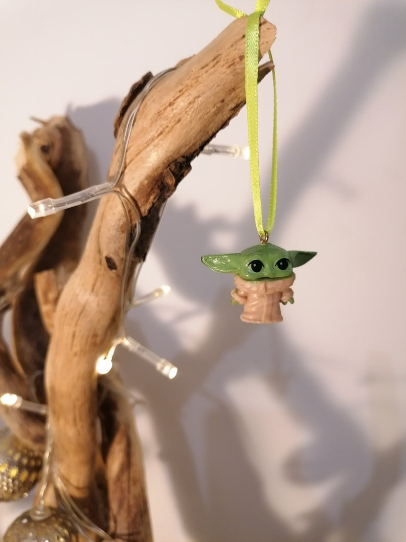 May include: A green and tan Baby Yoda ornament hanging from a light green ribbon. The ornament is suspended from a piece of driftwood, with a string of small white lights wrapped around it. The background is a plain white wall.