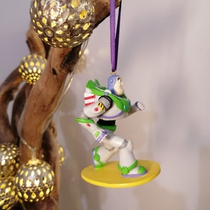 K&ouml;nnte beinhalten: Ein gr&uuml;ner und wei&szlig;er Plastik-Spielzeug-Anh&auml;nger von Buzz Lightyear aus der Toy Story-Filmreihe. Der Anh&auml;nger h&auml;ngt an einem lila Band und steht auf einem gelben Sockel.