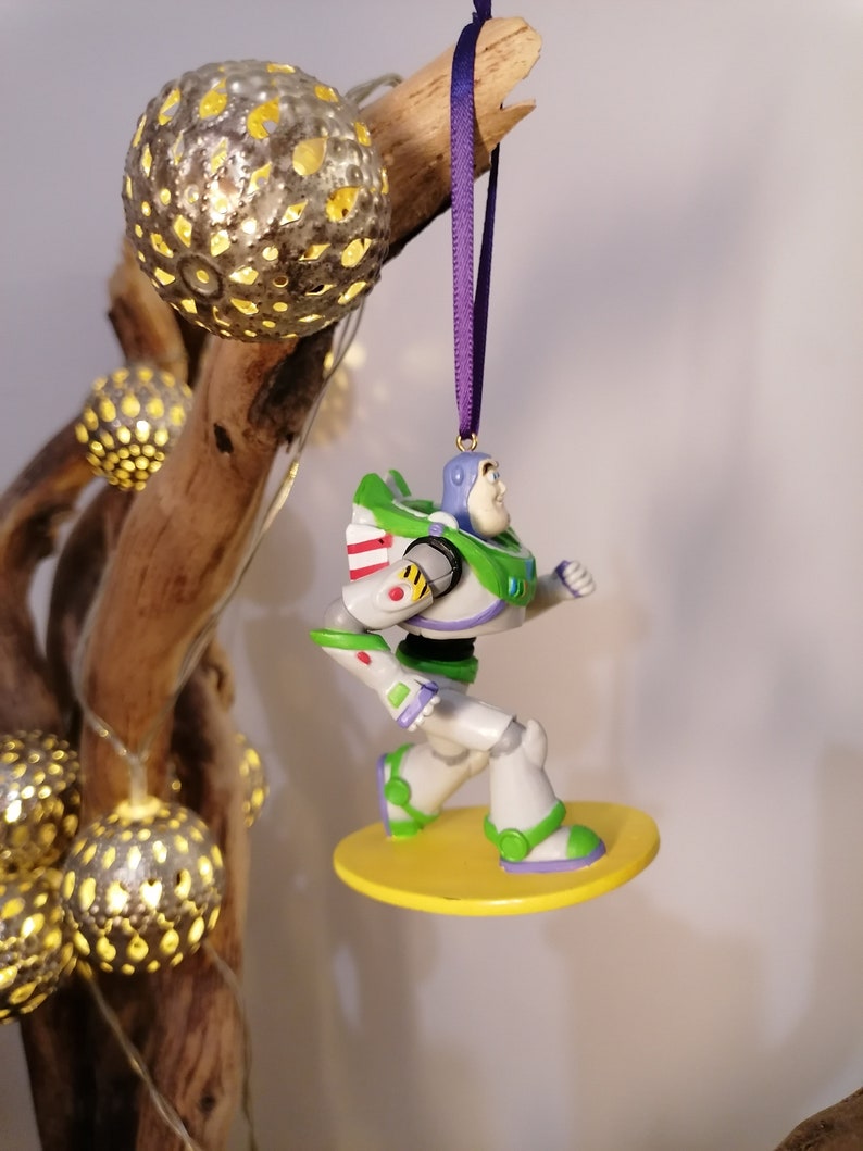 K&ouml;nnte beinhalten: Ein gr&uuml;ner und wei&szlig;er Plastik-Spielzeug-Anh&auml;nger von Buzz Lightyear aus der Toy Story-Filmreihe. Der Anh&auml;nger h&auml;ngt an einem lila Band und steht auf einem gelben Sockel.