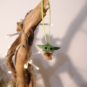 May include: A green and brown Yoda ornament hanging from a branch with a green ribbon. The ornament is a small, detailed figurine of the Star Wars character Yoda.
