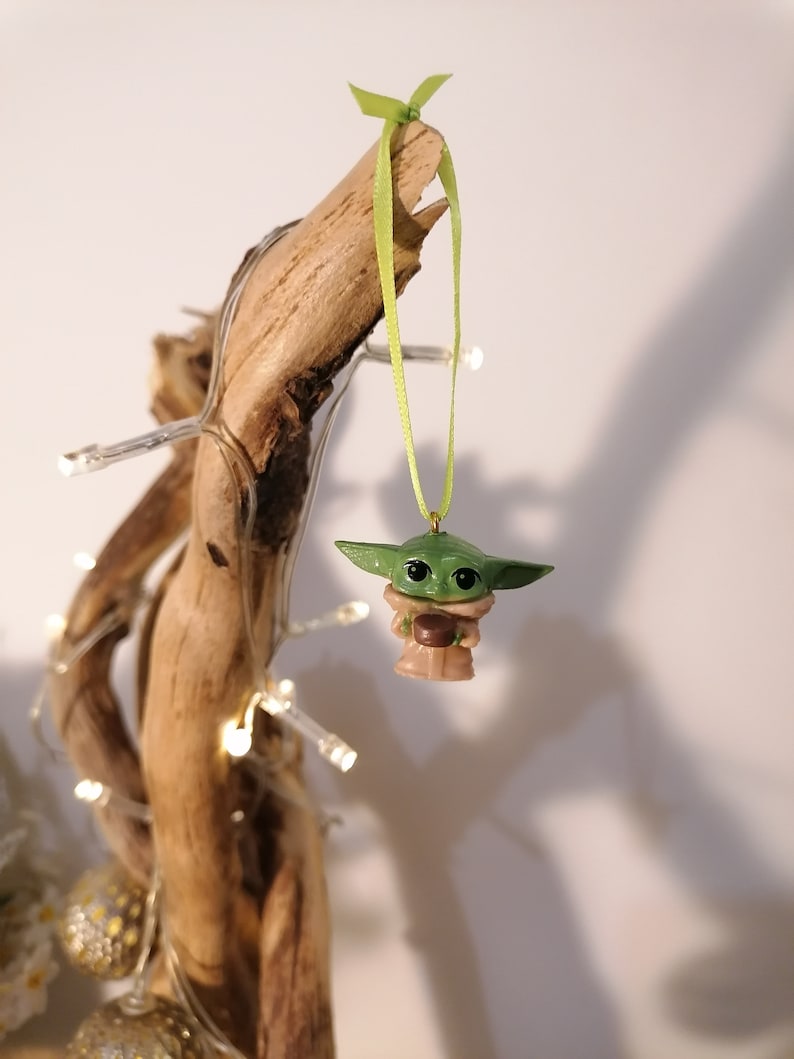 May include: A green and brown Yoda ornament hanging from a branch with a green ribbon. The ornament is a small, detailed figurine of the Star Wars character Yoda.