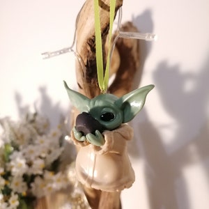 May include: A green and beige handmade ornament of a popular fictional character holding a small black object. The ornament is hanging from a green ribbon.