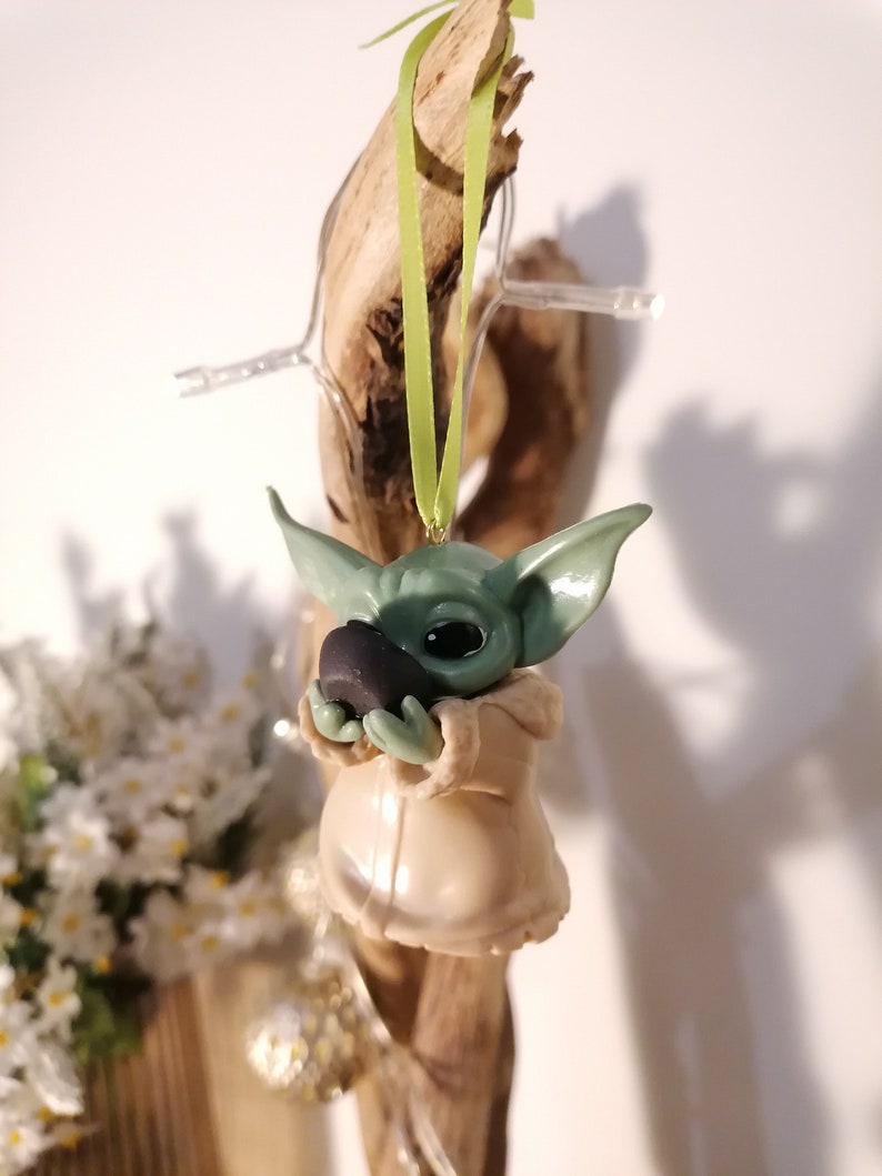 May include: A green and beige handmade ornament of a popular fictional character holding a small black object. The ornament is hanging from a green ribbon.