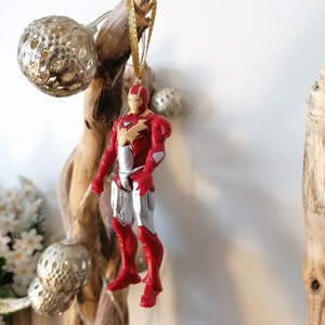 Pode incluir: Ornamento de figura de super-her&oacute;i Iron Man vermelho e prateado pendurado em uma fita dourada em um galho de madeira flutuante.