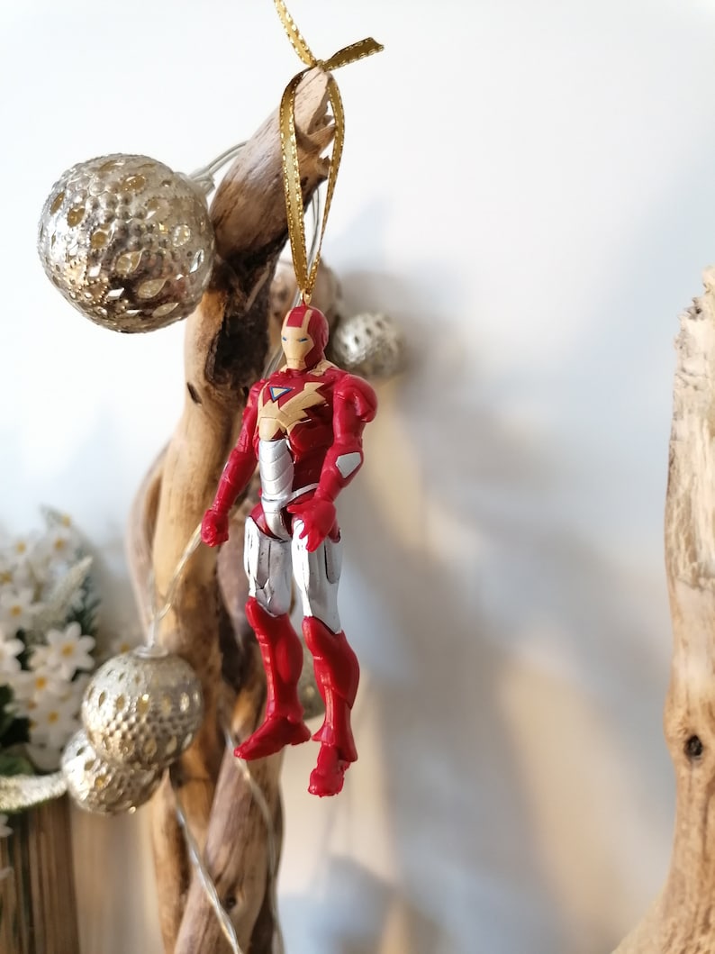 Pode incluir: Ornamento de figura de super-her&oacute;i Iron Man vermelho e prateado pendurado em uma fita dourada em um galho de madeira flutuante.