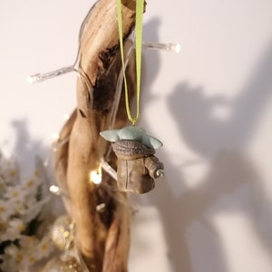Pu&ograve; includere: Una piccola statuina verde e marrone di un personaggio del franchise di Star Wars, appesa a un nastro verde su un pezzo di legno alla deriva.