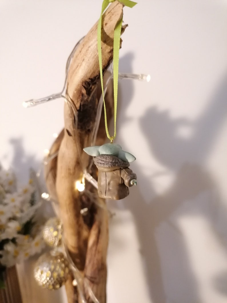 Pu&ograve; includere: Una piccola statuina verde e marrone di un personaggio del franchise di Star Wars, appesa a un nastro verde su un pezzo di legno alla deriva.