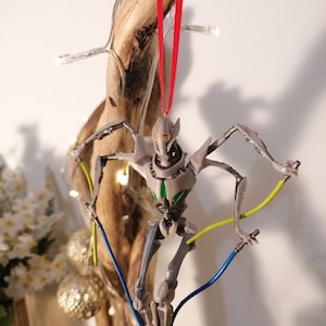 May include: A gray and green Star Wars General Grievous Christmas ornament with a red ribbon hanging from a branch. The ornament features a detailed sculpt of the character with lightsabers.