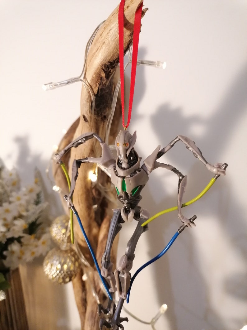 May include: A gray and green Star Wars General Grievous Christmas ornament with a red ribbon hanging from a branch. The ornament features a detailed sculpt of the character with lightsabers.