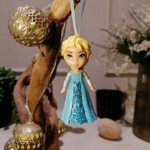 May include: A blue and silver glitter dress ornament of a Disney Frozen character, Elsa, with blonde hair and blue eyes. The ornament is hanging from a blue ribbon.