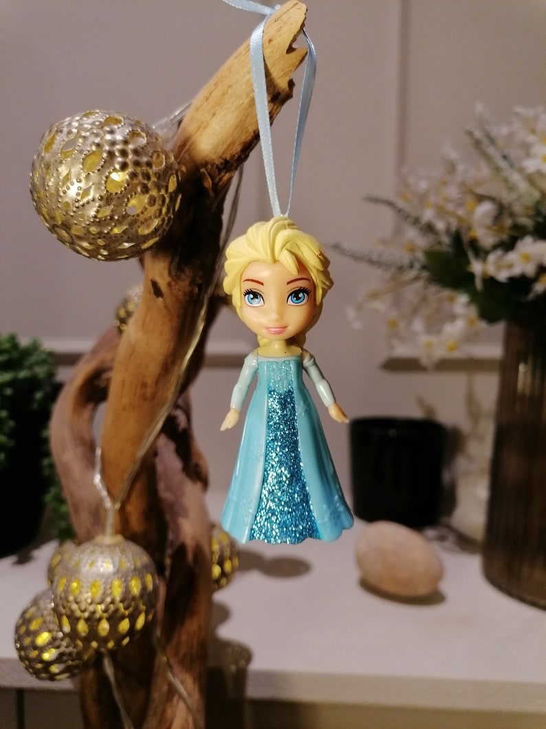 May include: A blue and silver glitter dress ornament of a Disney Frozen character, Elsa, with blonde hair and blue eyes. The ornament is hanging from a blue ribbon.