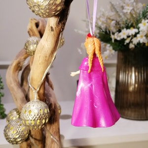 May include: A pink plastic doll with long orange hair in braids wearing a pink cape. The doll is hanging from a purple ribbon on a wooden branch with gold ornaments.
