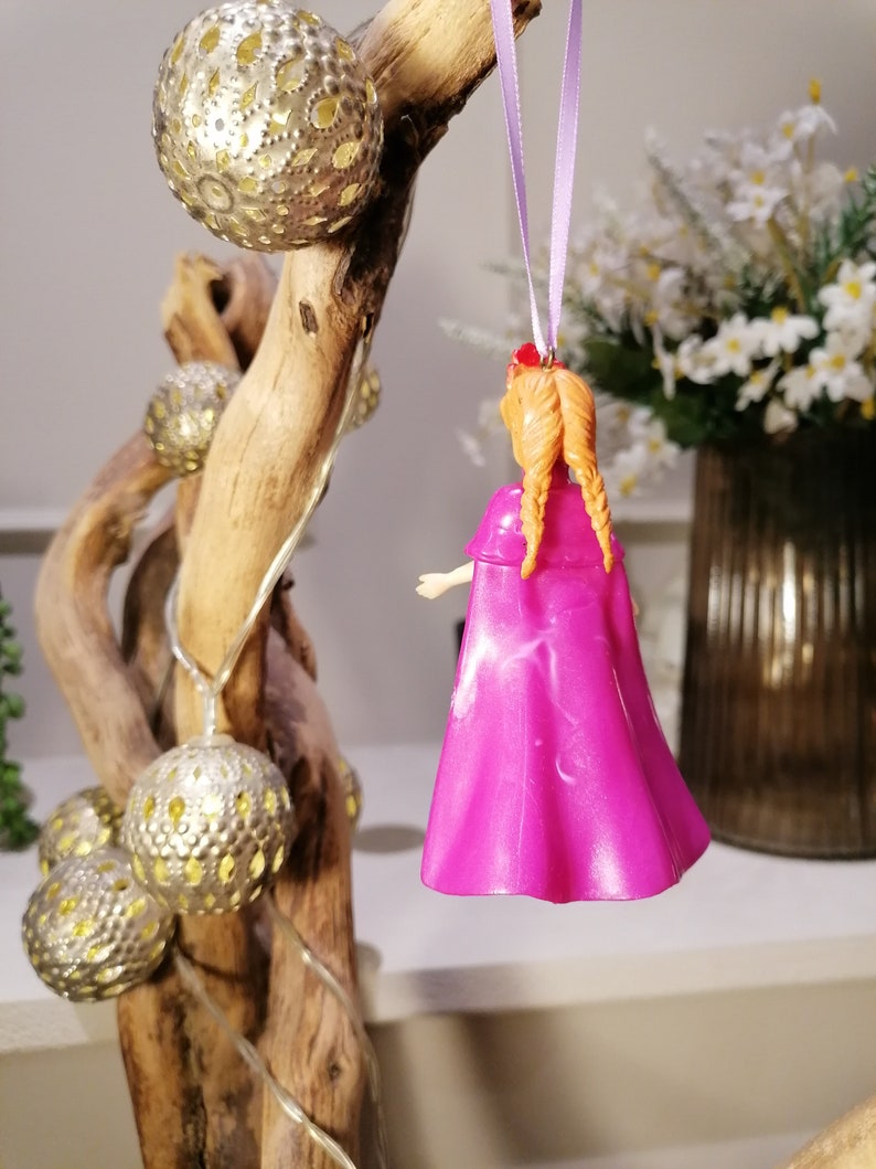 May include: A pink plastic doll with long orange hair in braids wearing a pink cape. The doll is hanging from a purple ribbon on a wooden branch with gold ornaments.