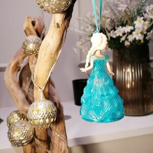 May include: A blue and white plastic figurine of a princess wearing a blue dress with a sparkly texture. The figurine is hanging from a blue ribbon on a branch with silver ornaments.