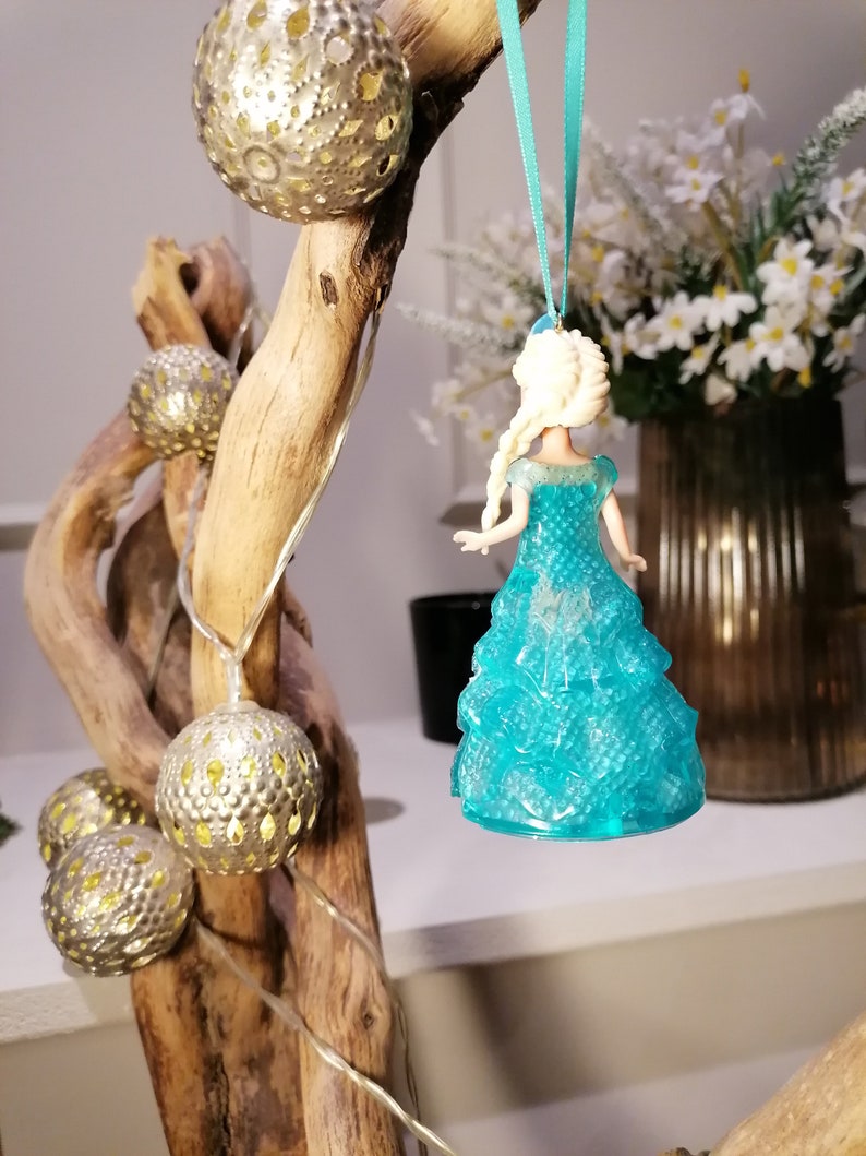 May include: A blue and white plastic figurine of a princess wearing a blue dress with a sparkly texture. The figurine is hanging from a blue ribbon on a branch with silver ornaments.