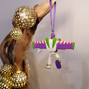 May include: A purple and white plastic toy astronaut ornament with wings hanging from a purple ribbon. The ornament is hanging from a branch with string lights.