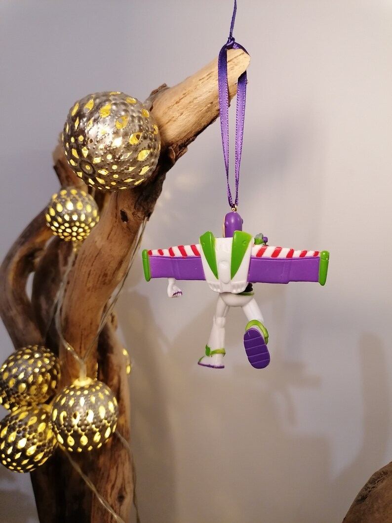 May include: A purple and white plastic toy astronaut ornament with wings hanging from a purple ribbon. The ornament is hanging from a branch with string lights.