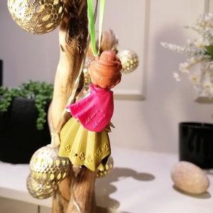 Pu&ograve; includere: Un piccolo ornamento di bambola di plastica con un vestito rosa e una gonna dorata. La bambola &egrave; appesa a un nastro verde su un ramo di legno.