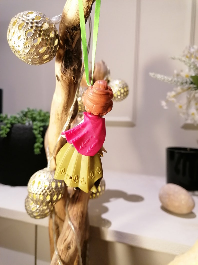 Pu&ograve; includere: Un piccolo ornamento di bambola di plastica con un vestito rosa e una gonna dorata. La bambola &egrave; appesa a un nastro verde su un ramo di legno.