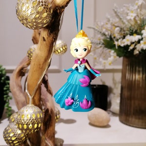 K&ouml;nnte beinhalten: Eine blaue und violette Plastikfigur einer Prinzessin mit blonden Haaren, die ein blaues Kleid und eine Krone tr&auml;gt. Die Figur h&auml;ngt an einem blauen Band.