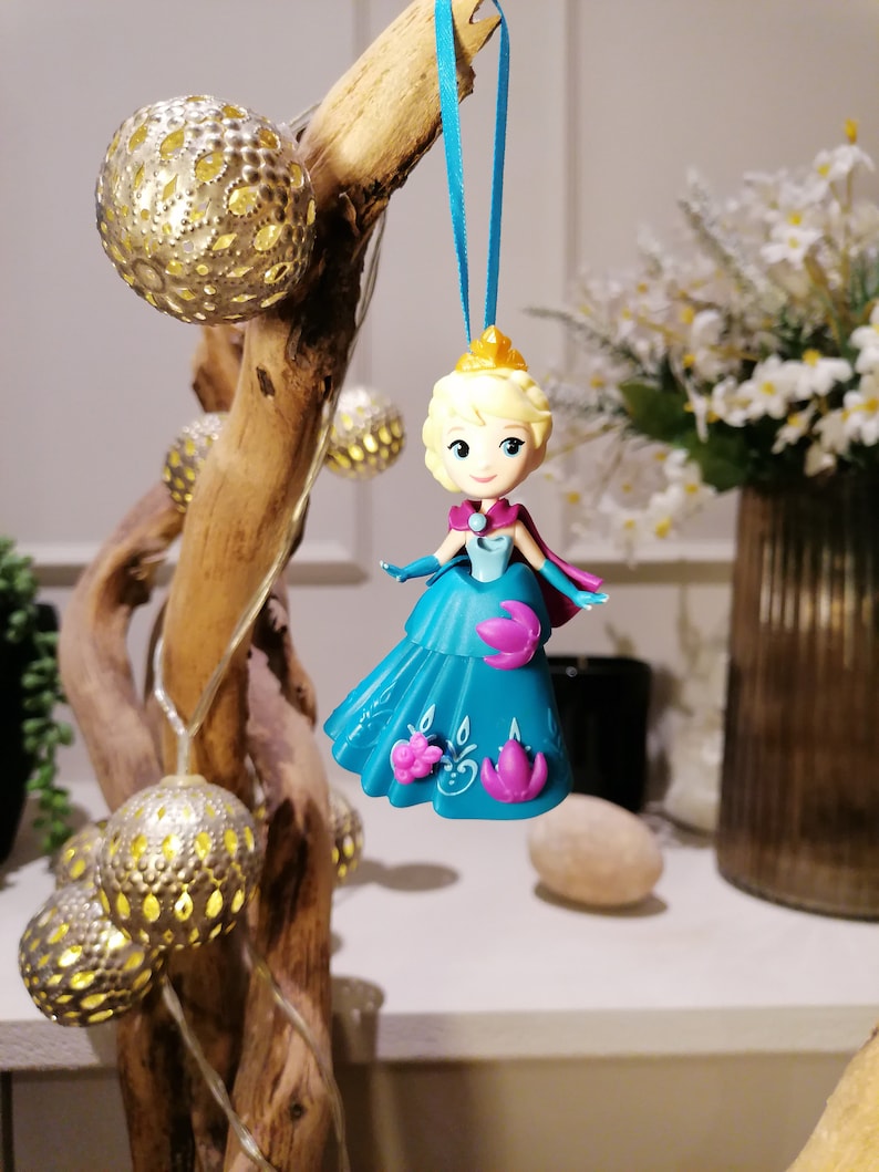 K&ouml;nnte beinhalten: Eine blaue und violette Plastikfigur einer Prinzessin mit blonden Haaren, die ein blaues Kleid und eine Krone tr&auml;gt. Die Figur h&auml;ngt an einem blauen Band.