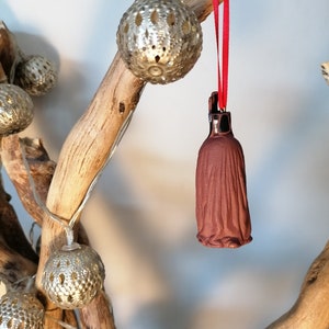 May include: A brown ceramic ornament shaped like a person wearing a long robe, hanging from a red ribbon on a branch with silver metallic ornaments.