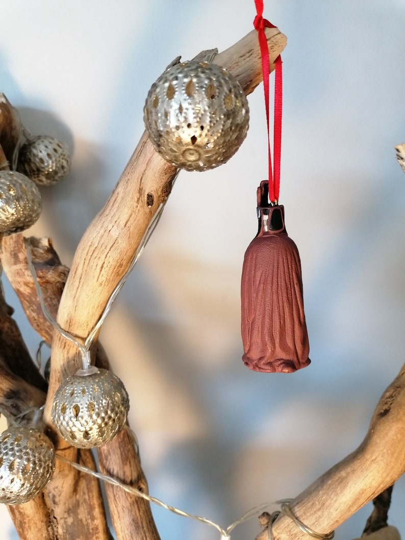 May include: A brown ceramic ornament shaped like a person wearing a long robe, hanging from a red ribbon on a branch with silver metallic ornaments.